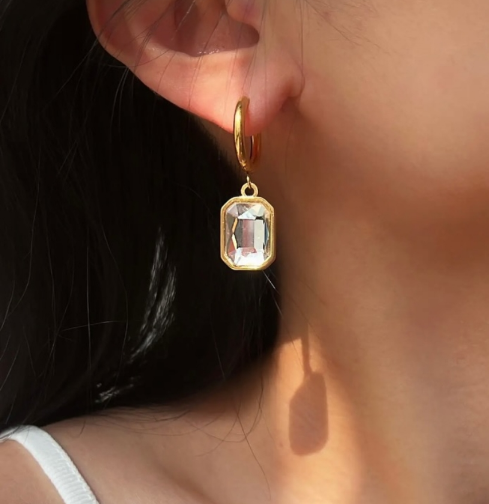 18k Gold-plated hoop earring with a clear rectangular zircon drop, shown worn on a models ear in a close-up.