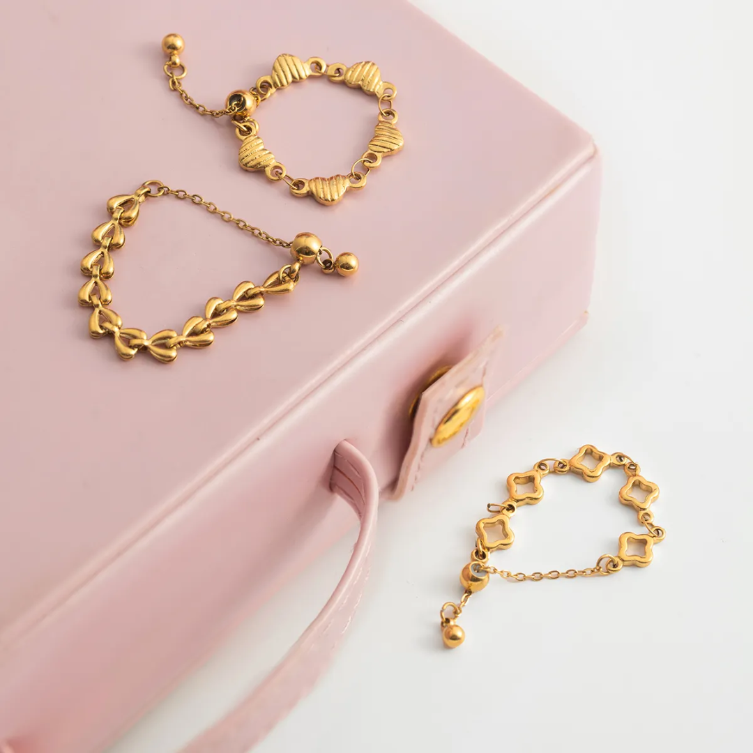 Three 18k Gold-plated chain rings with textured and geometric links are arranged on a light pink hard case against a white background.