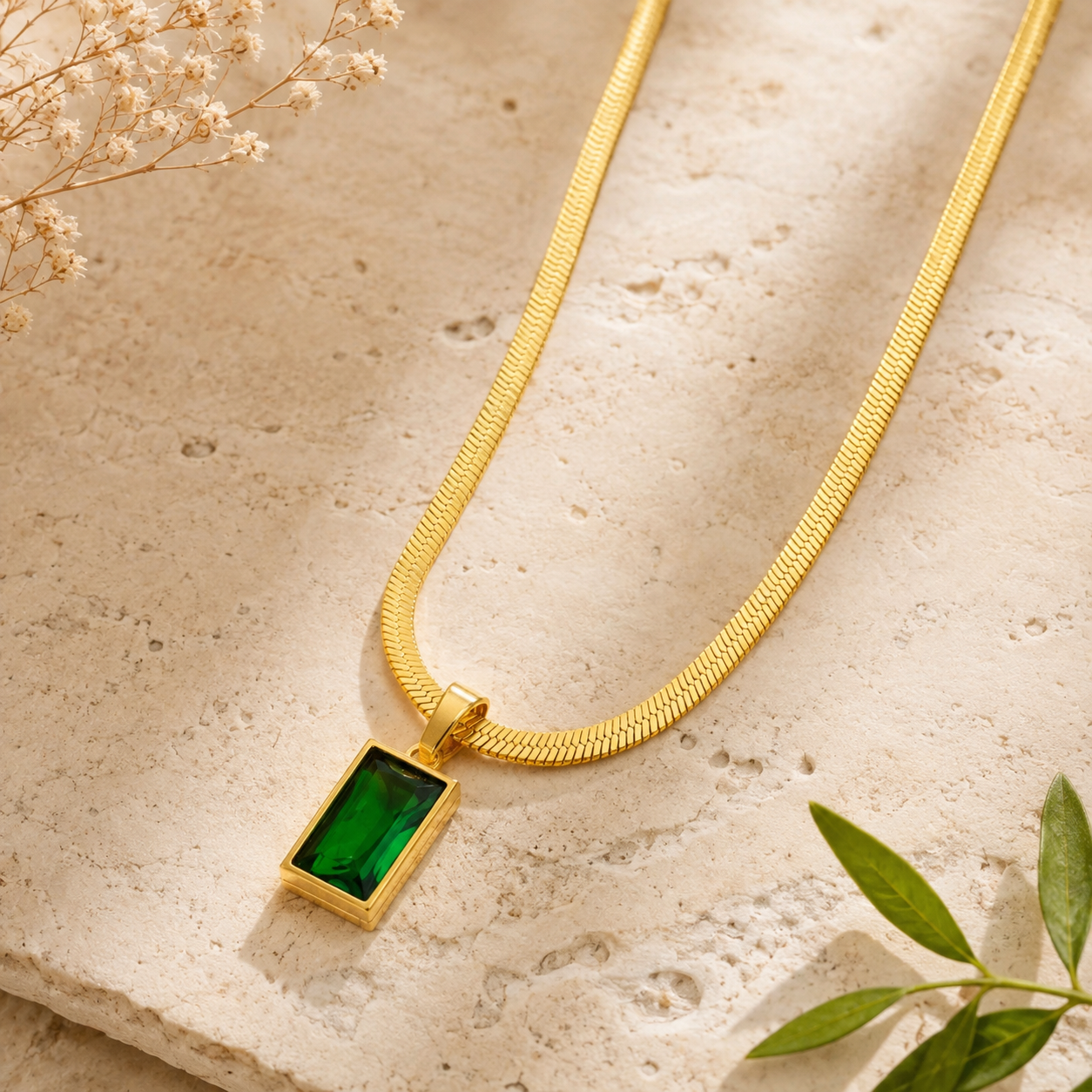 A gold snake chain necklace with a rectangular green gemstone pendant, styled on a textured beige stone surface with dried flowers and green leaves.