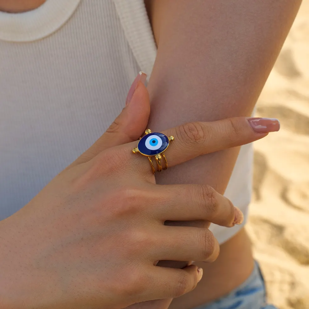 A 18k gold-plated ring with a blue and white evil eye design, worn on a person's finger in a beach setting.
