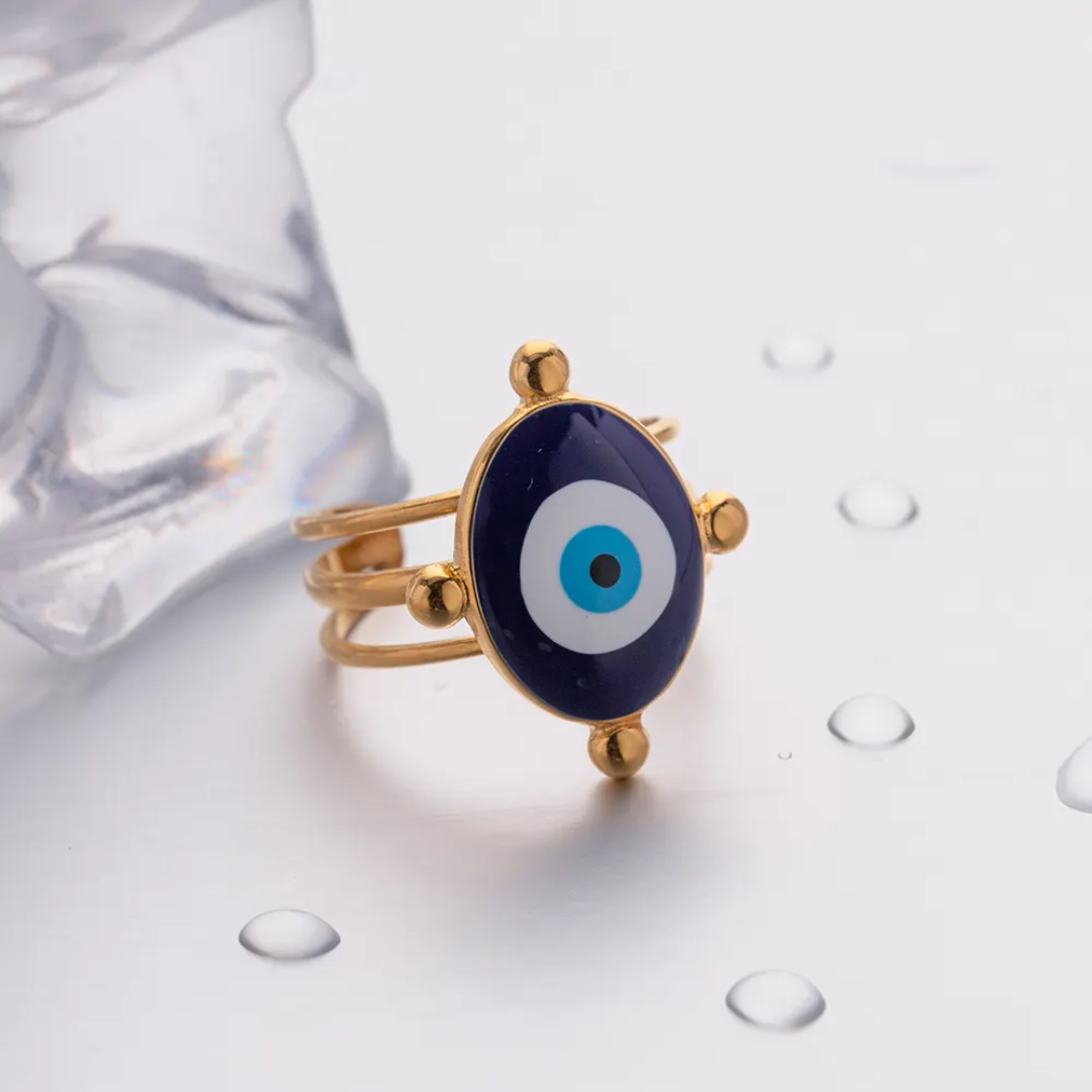 18k gold plated ring with an oval evil eye charm featuring navy blue, white, and light blue concentric circles, displayed on a white surface with water droplets and a clear textured prop.
