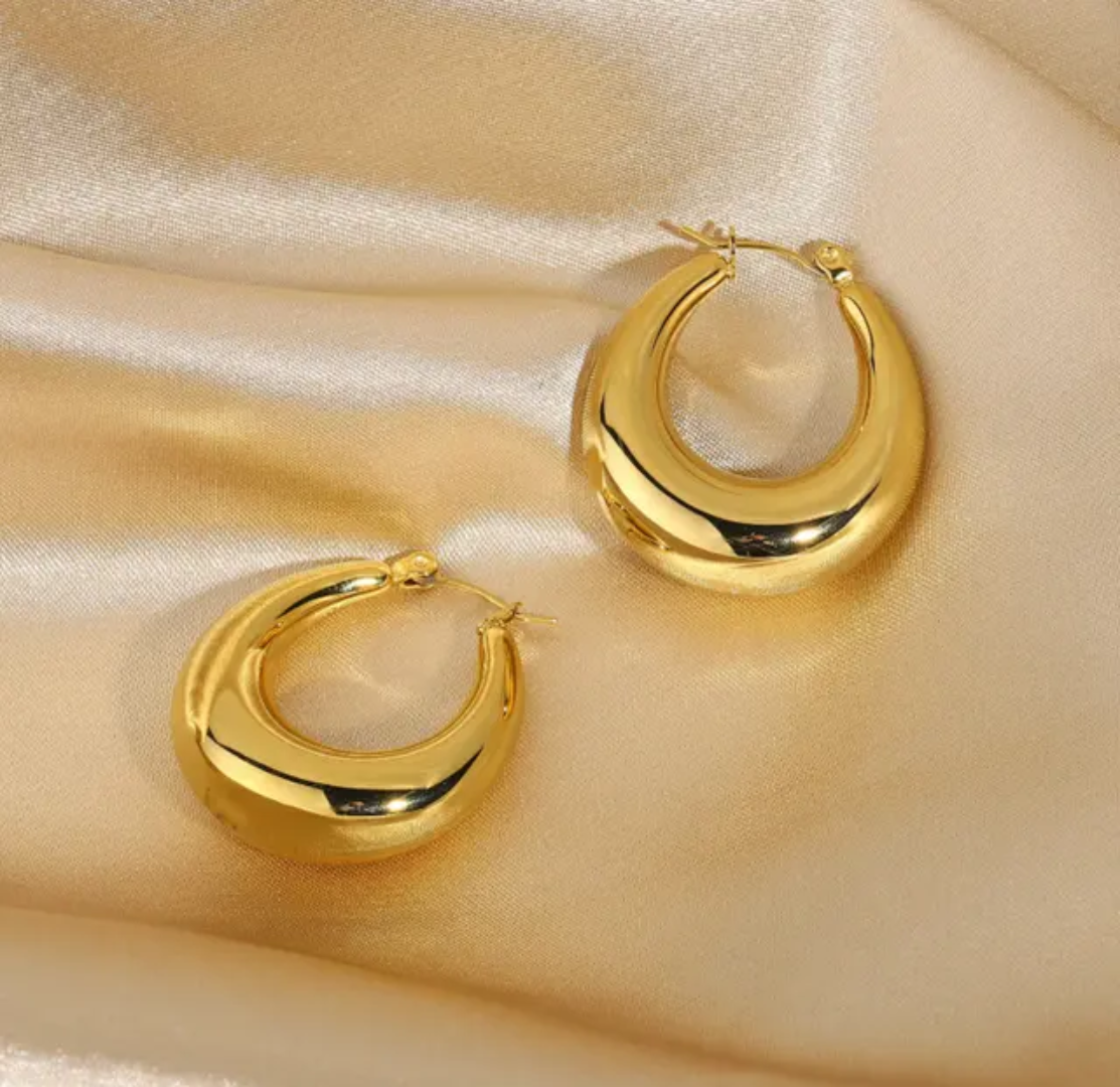 A pair of polished gold crescent hoop earrings displayed on a draped champagne-colored satin fabric background.