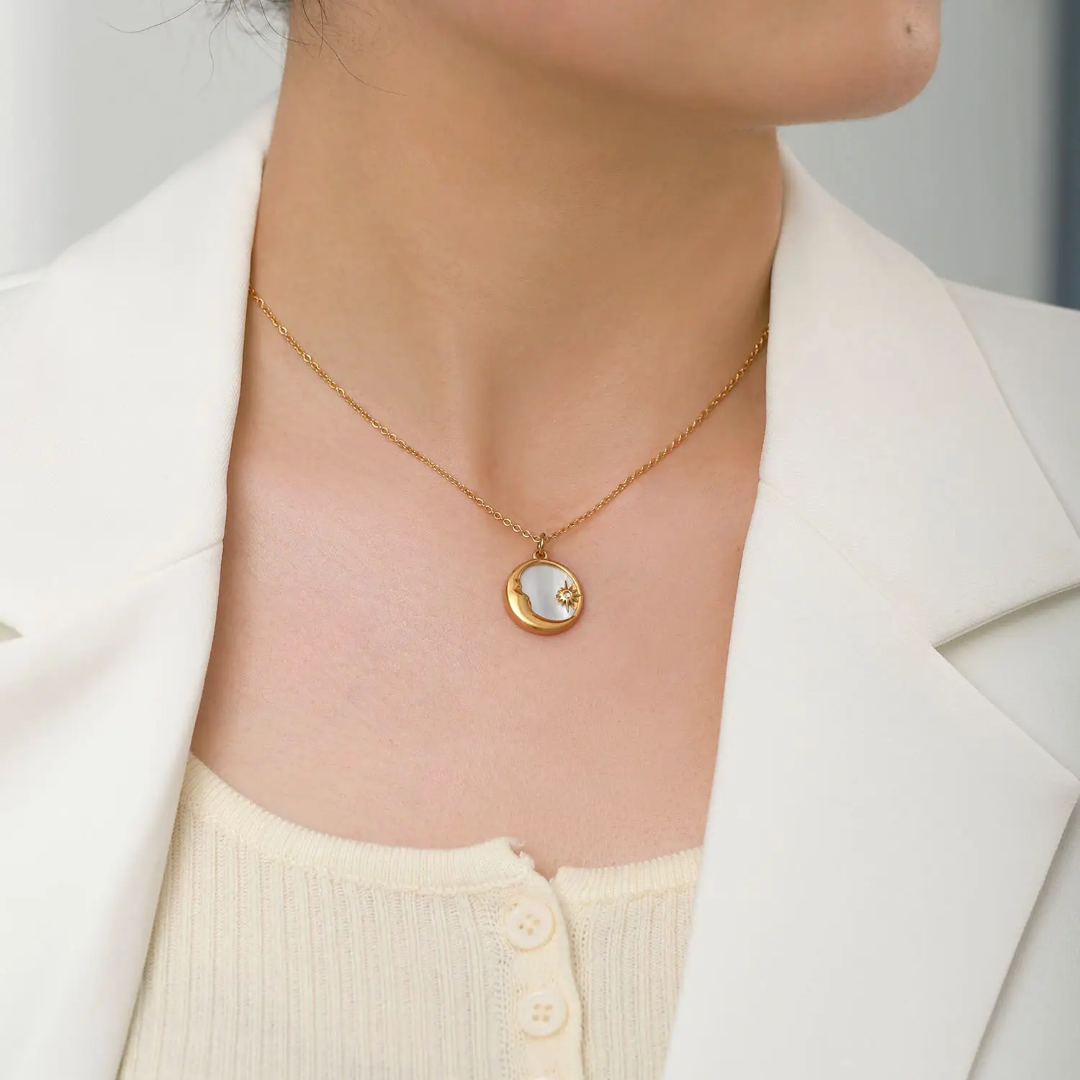 A close-up of a model wearing a 18k Gold-plated necklace with a small circular pendant featuring a crescent moon and star design, styled with a white blazer and cream top.
