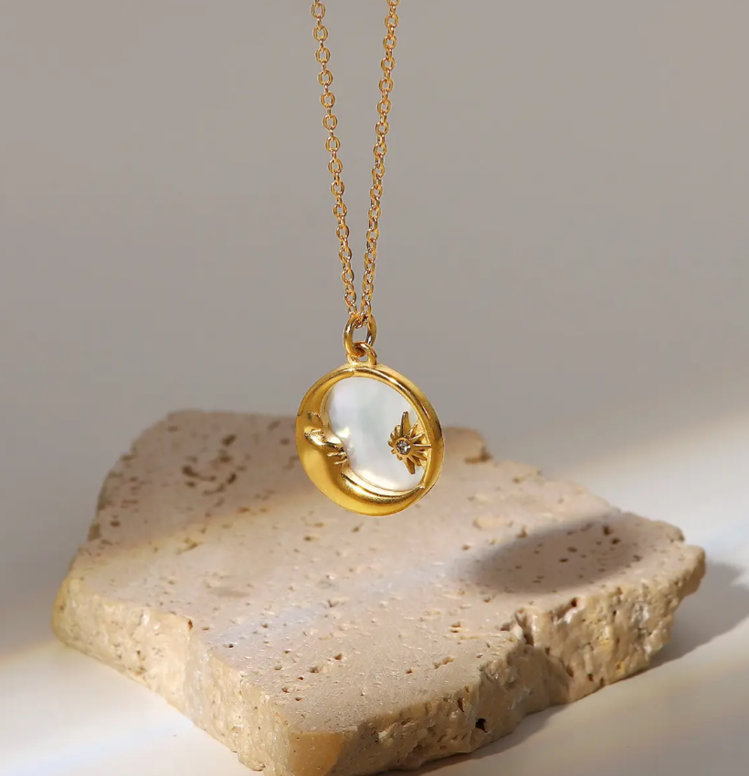 A gold crescent moon pendant necklace with a textured moon face and a small zircon star,  hanging on a gold chain above a beige stone prop against a neutral light gray background.