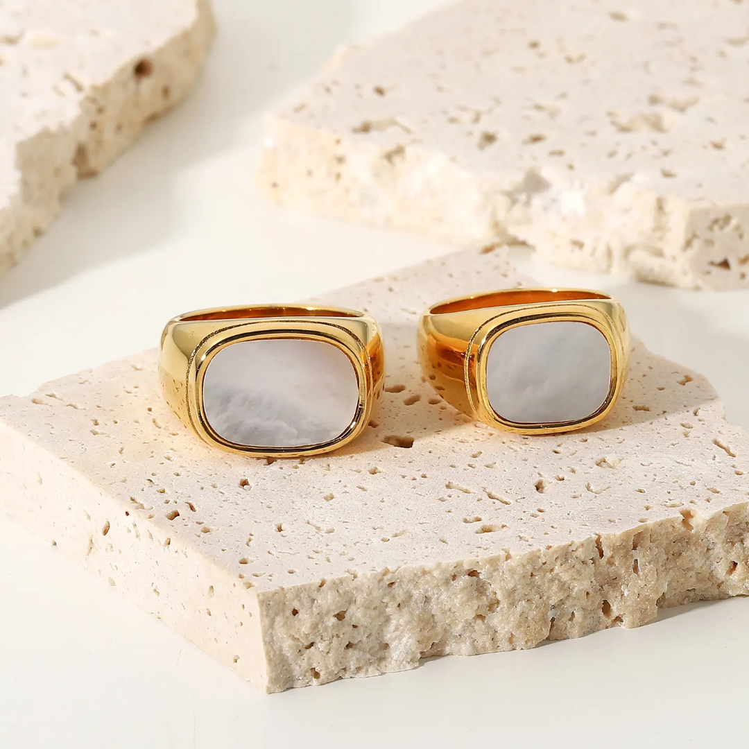 A pair of 18k gold plated signet rings with oval mother-of-pearl inlays, displayed on textured beige stone slabs against a clean white background.