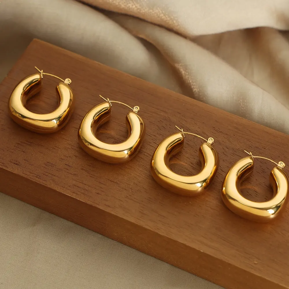 Four polished 18k gold plated chunky hoop earrings arranged on a wooden block with soft beige fabric in the background.