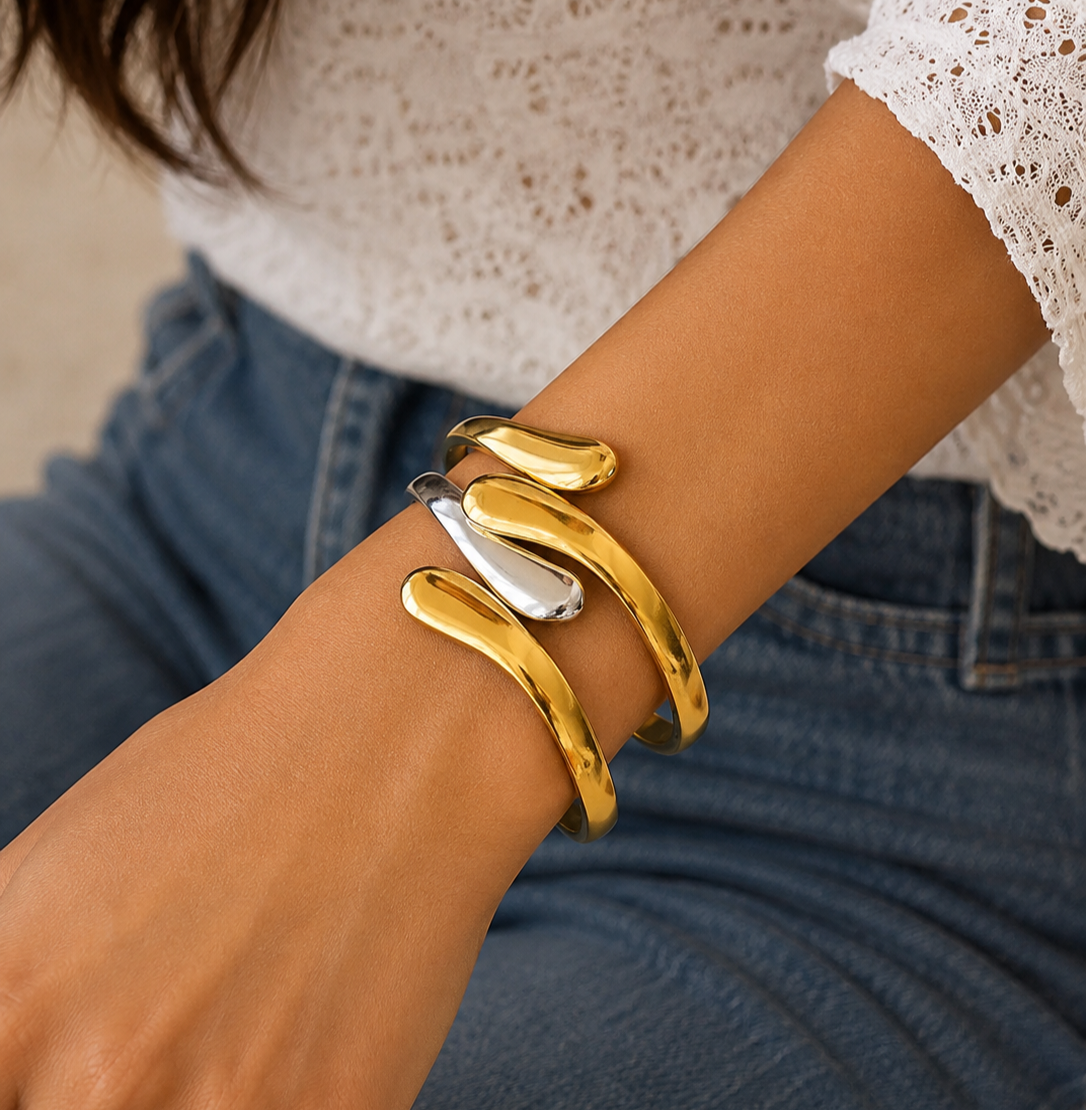 A close-up of a person's wrist wearing three open cuff bracelets, including two polished gold cuffs and one silver cuff, paired with a white lace top and blue jeans.