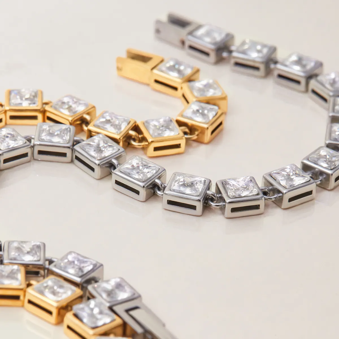 A close-up of a two square-link  tennis bracelet featuring clear zircons in silver-toned and gold-toned settings, arranged on a light cream background.