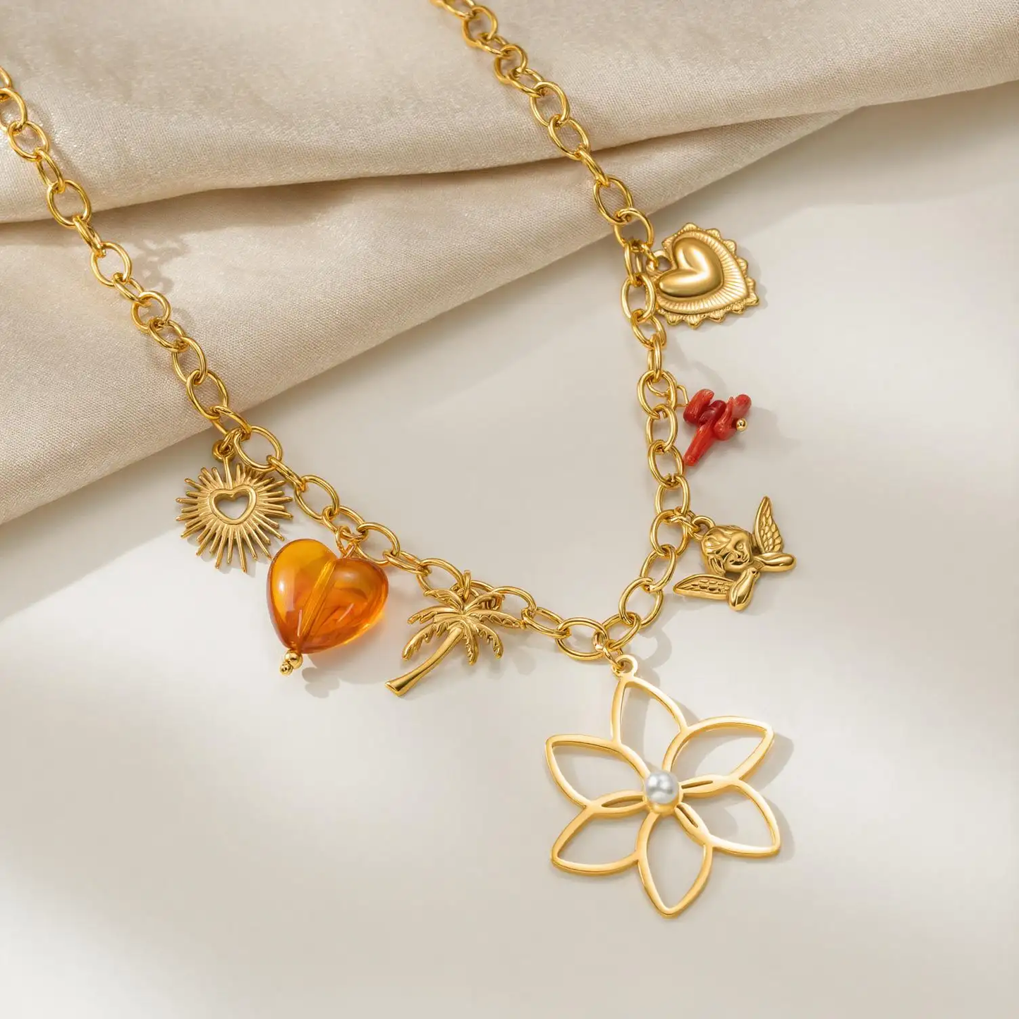 A 18k gold plated charm necklace displayed on a soft beige fabric background. The necklace features multiple charms, including a large gold-plated flower, a small red heart, a gold sun, a translucent amber heart, a palm tree, a bird, and a gold heart pendant.